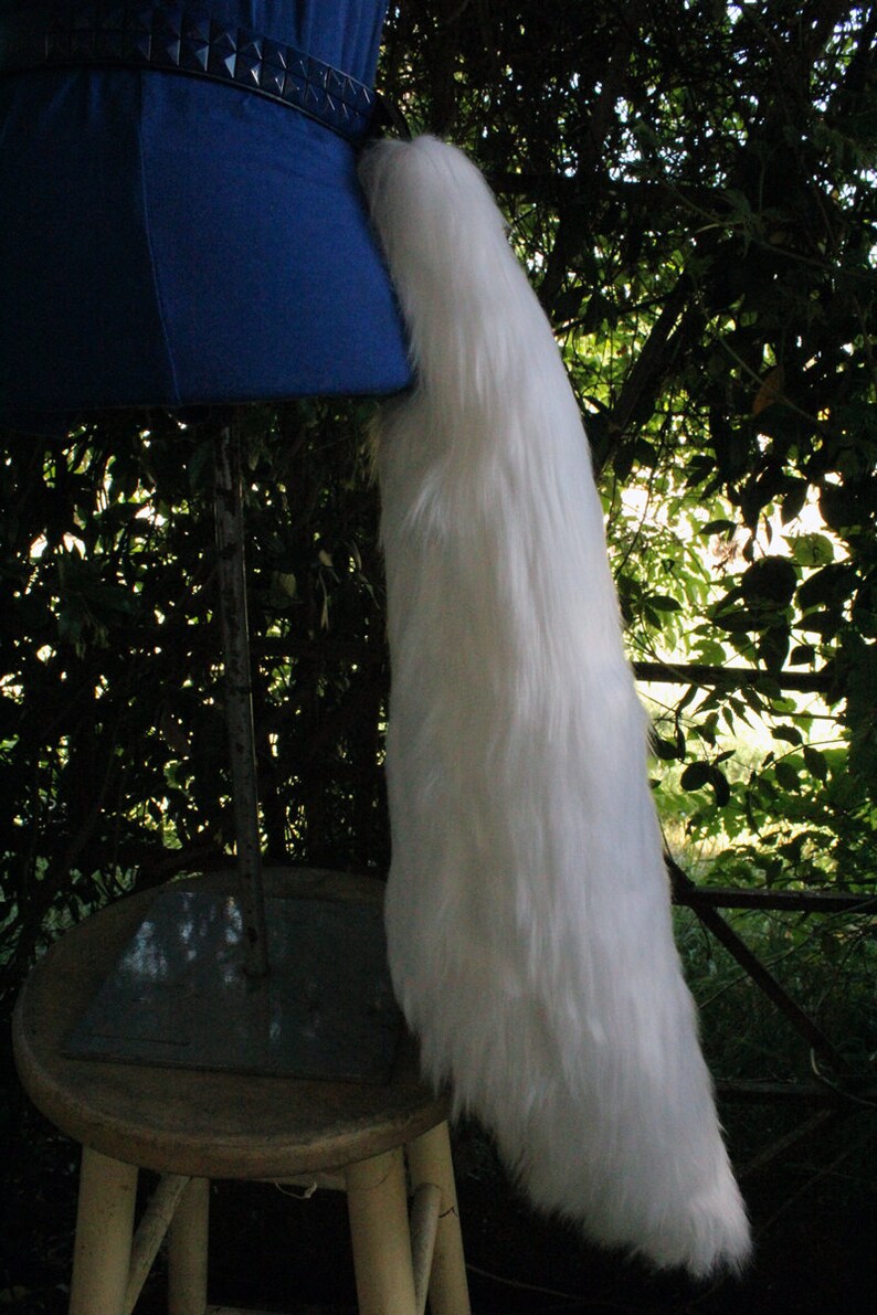 May include: A white fluffy tail prop, attached to a blue fabric base, is displayed on a wooden stool. The tail is long and full, with a soft, plush texture.