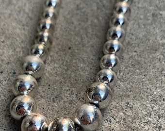 May include: A silver beaded necklace with a polished finish. The necklace features a series of round, reflective beads strung together. The beads vary slightly in size, creating a subtle visual texture. The necklace is displayed against a gray, textured surface.