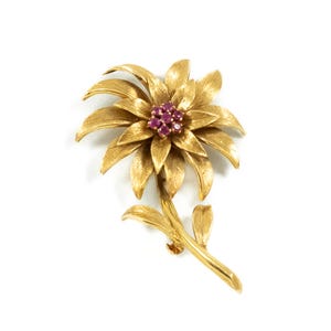 Può includere: Una spilla a forma di fiore in metallo dorato con petali testurizzati e un gruppo centrale di sette piccole pietre preziose rotonde color rosa intenso. Il design floreale presenta uno stelo curvo con due piccole foglie alla base del fiore.