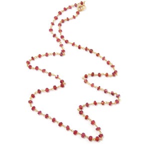 Puede incluir: Un collar largo con pequeñas piedras preciosas rojas facetadas ensartadas en una delicada cadena dorada. El collar tiene un cierre sencillo y se muestra sobre un fondo blanco.