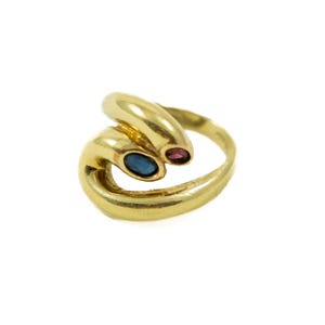 May include: A gold bypass ring featuring a smooth, polished band that curves around the finger. The ring is accented with two small, oval-shaped gemstones: one deep blue sapphire and one vibrant pink ruby, set at the ends of the bypass design.