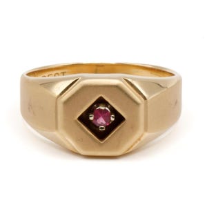 May include: A gold ring featuring a smooth, wide band with a central octagonal setting. A small, round, vibrant pinkish-red gemstone is prong-set in the middle of the octagon. The interior of the band shows a partial inscription.