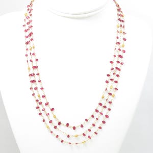 May include: A multi-strand necklace with ruby-colored beads and gold-colored accents. The necklace is displayed on a white mannequin bust. The beads are small and evenly spaced along the strands, creating a delicate and elegant look.