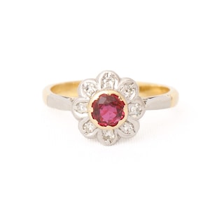 May include: A ring featuring a central round-cut red gemstone set in yellow gold, surrounded by a floral-shaped cluster of eight small white stones set in white metal, mounted on a polished yellow gold band with white metal shoulders.