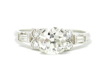 May include: A silver ring with a large, round, clear diamond center stone. The ring features smaller, clear diamonds and rectangular stones on either side of the center stone. The ring is set against a white background.