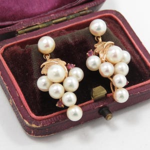 May include: A pair of gold-toned earrings featuring clusters of white pearls and small red gemstones. The earrings are designed to resemble grapes, with a leaf detail at the top. The earrings are displayed inside a vintage, burgundy jewelry box.