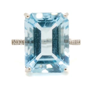 May include: A ring featuring a large, rectangular, emerald-cut light blue gemstone set in a silver-toned metal prong setting. The band of the ring displays a textured, repeating pattern along its visible sides, extending from the central stone.