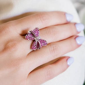 May include: A silver ring with a butterfly design. The butterfly is made of small, bright pink gemstones and has a small diamond in the center. The ring is on a hand with light purple nail polish.