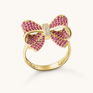 May include: A gold-colored ring in the shape of a bow. The bow is encrusted with small, round, pink gemstones. The center of the bow features a band of clear gemstones. The ring band is smooth and polished.