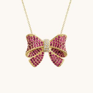 Puede incluir: Un collar dorado con un colgante de lazo. El lazo está adornado con pequeñas piedras preciosas rosas redondas y una banda central de piedras claras y brillantes. La cadena del collar es delicada y dorada.