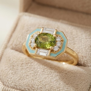 May include: A gold ring with a light green oval gemstone at the center, surrounded by small white diamonds and rectangular clear stones. The ring has a light blue enamel border and is set in a gold band. The ring is displayed in a beige ring box.