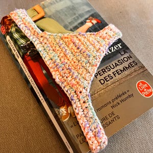 Puede incluir: Un marcapáginas de ganchillo hecho a mano, de colores, con forma de tanga, descansa sobre un libro titulado "LA PERSUASION DES FEMMES". El marcapáginas presenta una mezcla de colores pastel, como rosa, azul y amarillo. El libro está en francés.