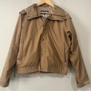 May include: A tan jacket with a hood, button and zipper closure, and two front pockets. The jacket has a ribbed hem and cuffs. The brand name "Windy Pass" is visible on the label. The jacket is hanging on a wooden hanger.