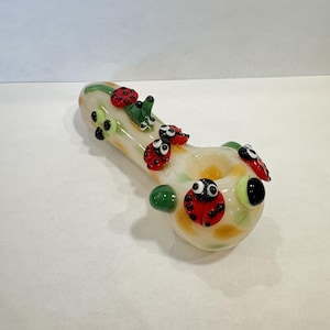 May include: A clear glass pipe featuring a cream-colored base with yellow and green accents. The pipe is decorated with several small, three-dimensional glass ladybugs in red and black, along with small green glass caterpillar-like figures.