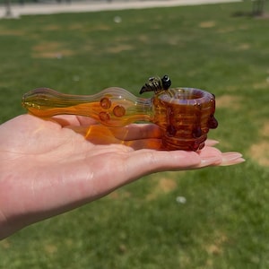 May include: A translucent amber glass pipe featuring a small, detailed bumblebee figurine attached to the side of the bowl. The pipe has a smooth, rounded stem with three small glass dots and a textured, cylindrical bowl with raised glass accents.
