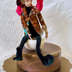 Op de afbeelding: Een grillige pop met een aapachtig gezicht, gekleed in een jasje met patroon, een rode hoed en een groene fluwelen broek. De pop zit in een witte kom op een decoratieve houten en messing doos. De pop heeft een speelse uitstraling.