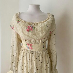 May include: A vintage cream-colored dress with a floral print and black polka dots. The dress has a ruffled neckline and long sleeves.