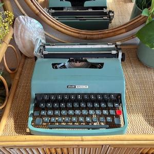 Puede incluir: Una máquina de escribir Olivetti Lettera 22 turquesa vintage con teclas negras y una tecla de retorno roja. La máquina de escribir está sobre una superficie tejida, con el reflejo de otra máquina de escribir en un espejo.