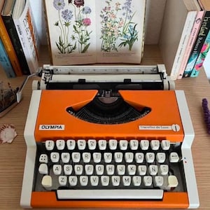 May include: A vintage orange Olympia Traveller de Luxe typewriter with white keys. The typewriter is set on a wooden surface, with books and floral prints in the background. The text "Traveller de Luxe" is visible.