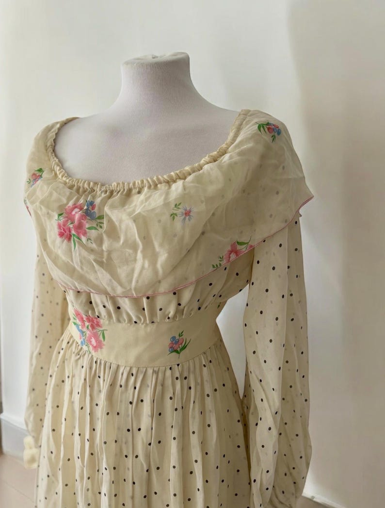 May include: A vintage cream-colored dress with a floral print and black polka dots. The dress has a ruffled neckline and long sleeves.