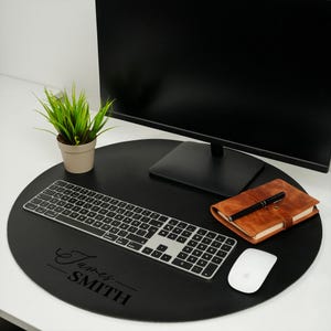 Puede incluir: Una alfombrilla de escritorio redonda negra con una inscripción personalizada "James Smith". Un teclado, un ratón y una libreta de cuero marrón están colocados sobre la alfombrilla.