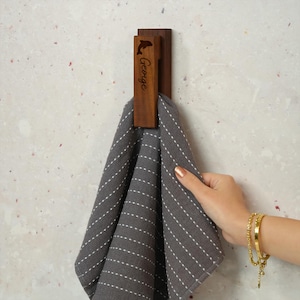 Könnte beinhalten: Ein hölzerner Handtuchhaken mit einer Delfin-Silhouette und dem eingravierten Namen "George". Ein graues Handtuch mit weißen Streifen hängt an dem Haken. Der Haken ist an einer hellen Wand befestigt.