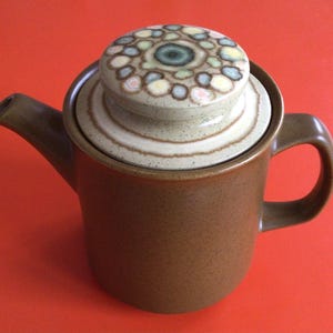 May include: A brown ceramic coffee pot with a lid featuring a colourful geometric design. The lid has a white rim and a brown base.