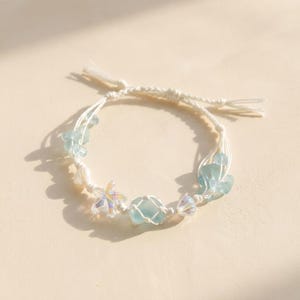 May include: A delicate white cord bracelet decorated with light blue and iridescent beads, including starfish and seashell shapes. The bracelet features a central, light blue, sea glass-like bead.