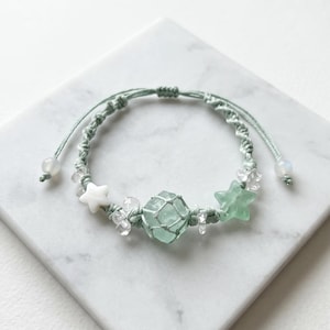 May include: A handmade macrame bracelet in shades of green, featuring a central seafoam-colored stone encased in a net. The bracelet is adorned with white star and clear crystal beads, and adjustable cords.