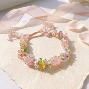 May include: A delicate pink bracelet featuring large, faceted pink stones interspersed with small, colorful floral accents. The bracelet is adorned with tiny gold-colored beads and a matching pink ribbon.
