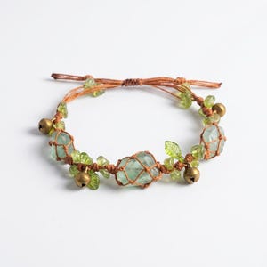 Puede incluir: Una pulsera de macramé hecha a mano con piedras preciosas de color verde claro, pequeñas campanas de latón y detalles de hojas verdes. La pulsera está ensartada en un cordón marrón y tiene un cierre ajustable.