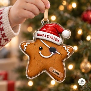 May include: A star-shaped gingerbread ornament featuring a red Santa hat with the text WHAT A YEAR 2026, a black eye patch, and a white icing smile. A tan bandage on the lower right side displays the name SOPHIA in black capital letters.