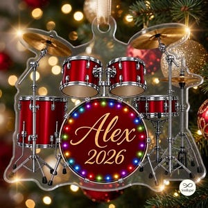 Op de afbeelding: Een doorzichtig acryl kerstornament in de vorm van een drumstel met rode trommels en gouden bekkens. De bassdrum heeft een cirkelvormig ontwerp met gekleurde lichtjes langs de rand en de tekst Alex 2026 in gouden letters en cijfers.