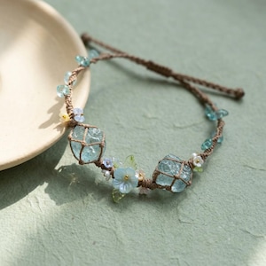Puede incluir: Una pulsera de macramé hecha a mano con dos grandes piedras de aguamarina azul claro. La pulsera está adornada con pequeñas cuentas azules y transparentes, diminutas perlas blancas y delicados detalles de flores azules. El cordón de macramé es de color marrón natural.