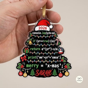 Op de afbeelding: Een doorzichtig, boomvormig acryl ornament met een zwarte achtergrond, kleurrijke kerstverlichting en programmeercode. Het ontwerp bevat een rode kerstmuts aan de bovenkant, het jaartal 2026 en de naam SARAH in rode letters onderaan.