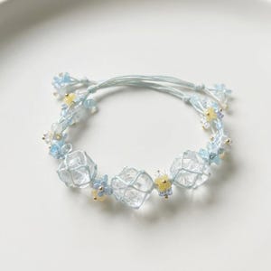 Puede incluir: Una delicada pulsera con cuentas claras y facetadas, encerradas en una red azul claro. La pulsera está adornada con pequeños acentos florales azules y amarillos y ensartada en un cordón azul claro. El diseño se encuentra sobre un fondo blanco.