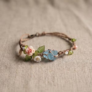May include: A handcrafted macrame bracelet with a light blue crystal, pink rose, green leaves, and small white flowers. The bracelet is decorated with small green and pink beads. The bracelet is made with a brown cord.