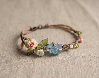 May include: A handmade macrame bracelet featuring a light blue crystal, pink rose, green leaves, and small white flowers. The bracelet is adorned with small green and pink beads. The bracelet is made with a brown cord.