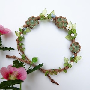 May include: A handcrafted bracelet featuring three green crystal orbs encased in a macrame net design. The bracelet is adorned with green leaves, tiny white and blue flowers, and is made with brown twine. The bracelet is set against a white background with pink flowers.