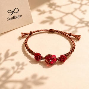 May include: A brown macrame bracelet featuring three red accents: two rose-shaped beads and a heart-shaped bead. The bracelet is displayed on a light-colored surface. The brand name "Soullogue" is visible on a card.