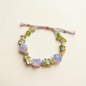 May include: A delicate bracelet featuring three large, light purple, rounded beads. The bracelet is adorned with small white, blue, and green floral accents and green beads. The bracelet is strung on a pastel pink, blue, and orange cord.