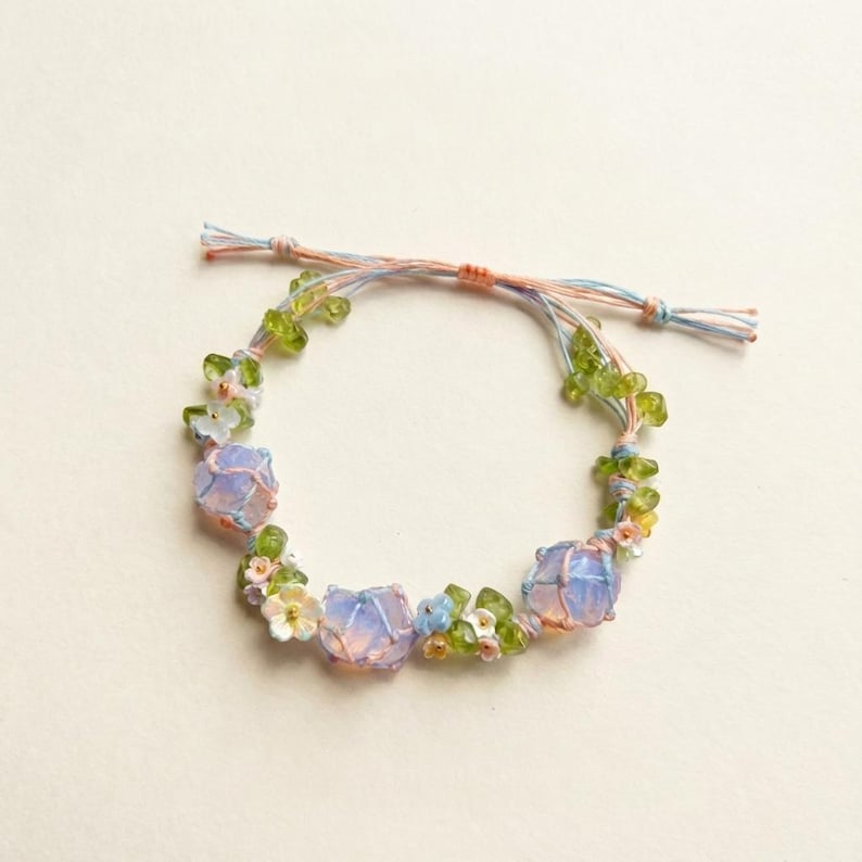 May include: A delicate bracelet featuring three large, light purple, rounded beads. The bracelet is adorned with small white, blue, and green floral accents and green beads. The bracelet is strung on a pastel pink, blue, and orange cord.