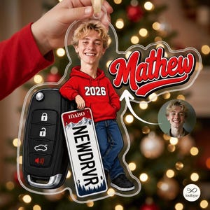 May include: A clear acrylic ornament featuring a cartoon illustration of a blonde person in a red hoodie with the text 2026, leaning against a black car key fob and an Idaho license plate that reads NEWDRVR, with the name Mathew in red script.