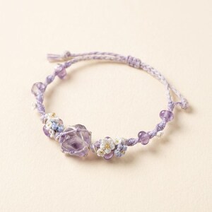 May include: A handmade lavender bracelet featuring a large amethyst crystal, small purple beads, and delicate floral accents. The adjustable cord bracelet is a light purple color and is designed for a comfortable fit.