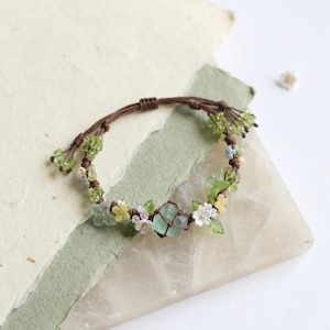 May include: A handmade bracelet featuring a brown cord adorned with green, yellow, and white floral accents. The bracelet incorporates small green beads and larger, light green crystal-like stones, creating a delicate, nature-inspired design.