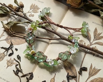 May include: A handmade bracelet featuring green crystal stones encased in a brown net, accented with white and green floral beads. The bracelet is displayed on an open book with botanical illustrations and dried flowers.
