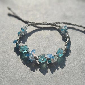 May include: A handmade bracelet featuring light blue and clear crystal beads, woven with a gray cord. The bracelet has a delicate design with a mix of different sized beads and small floral accents. The bracelet is shown on a gray surface.