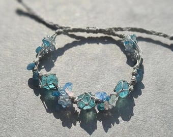 May include: A handmade bracelet featuring light blue and clear crystal beads, woven with a gray cord. The bracelet has a delicate design with a mix of different sized beads and small floral accents. The bracelet is shown on a gray surface.
