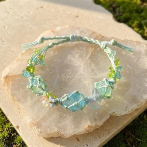 May include: A handmade macrame bracelet featuring light blue and green accents. The bracelet incorporates square-shaped, light blue glass beads, small green beads, and delicate floral embellishments. The bracelet is displayed on a light-colored stone surface.