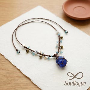 May include: A handcrafted necklace featuring a dark brown cord with a blue gemstone pendant. The necklace is adorned with small blue and white beads, along with tiny gold-colored bells. The necklace rests on a white, textured surface.